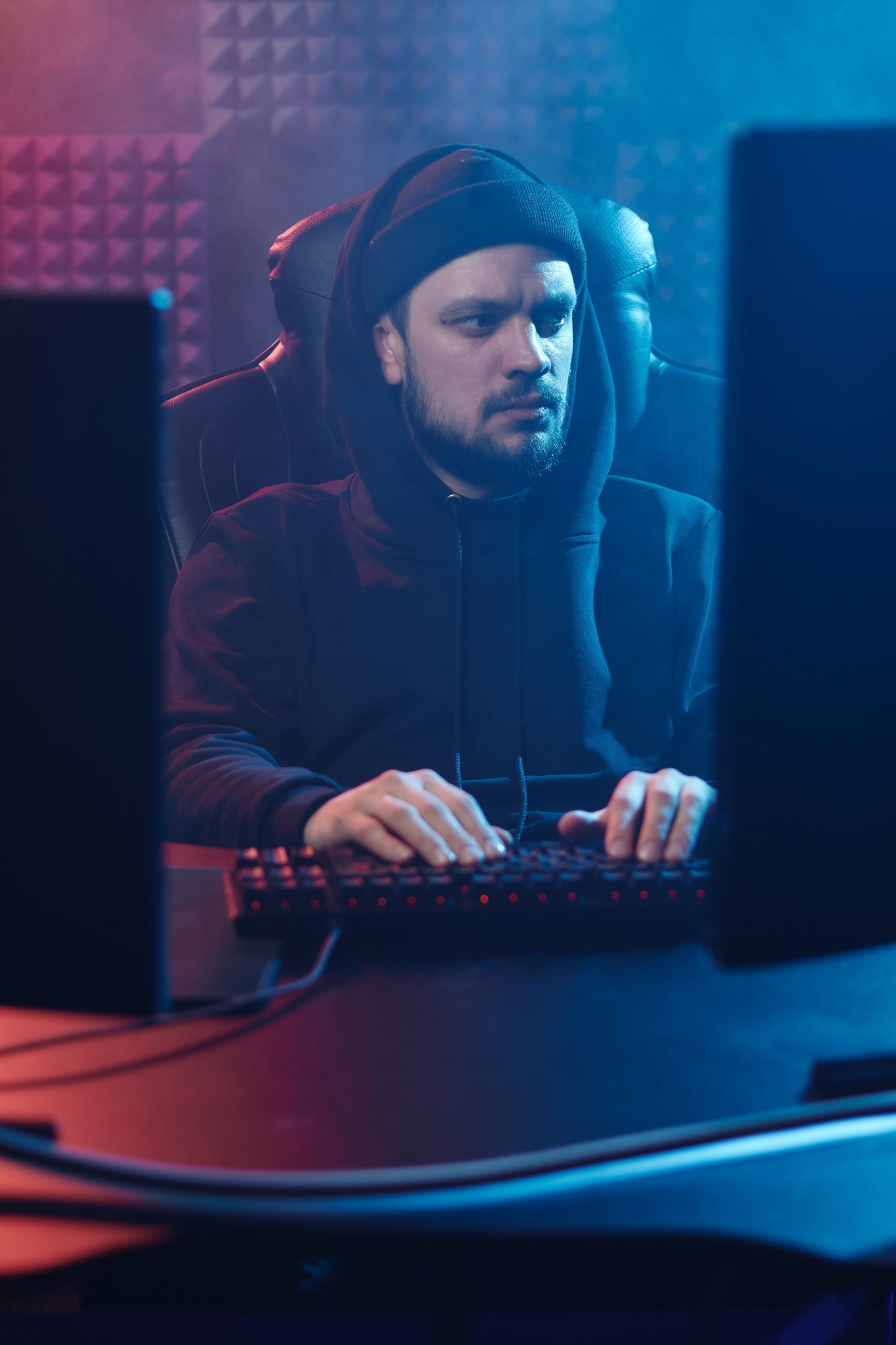 Guy with a goatee in a black hoodie looks at one of two computer monitors; his hands on a keyboard; dark environment with soundproofing